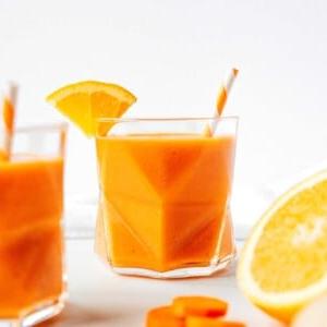 Ein geometrisches Glas Karotten-Smoothie, gemixt mit leuchtender Orange, garniert mit einem frischen Orangenkeil und Strohhalm. Geschnittene Karotte und eine halbe Orange liegen verlockend im Vordergrund.