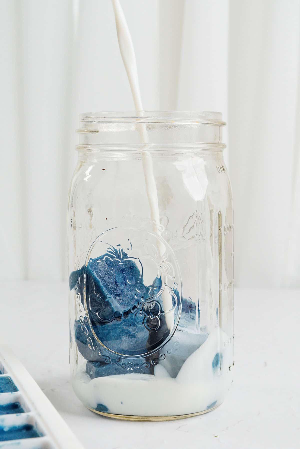 Smoothie-Würfel (Evive-Kopie) 9 Milch wird über blaue Smoothie-Würfel in einem Glas gegossen.