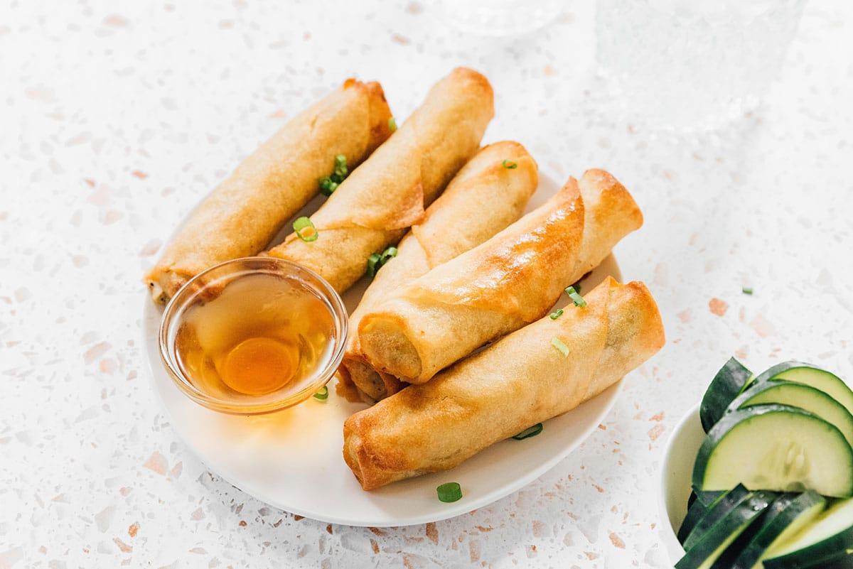 Die knusprigsten tiefgekühlten Frühlingsrollen aus der Heißluftfritteuse 9 Heißluftfrittierte Frühlingsrollen auf einem Teller neben Gurken und einer Dip-Sauce.