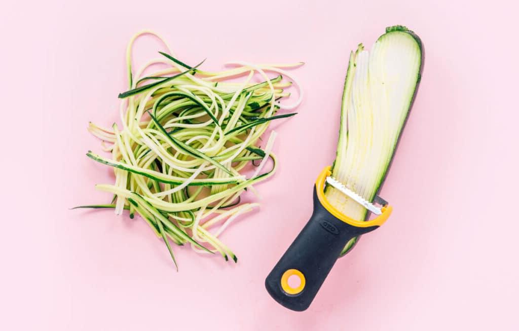 Zucchininudeln zubereiten 9 Ein Stapel engelshaariger Zucchini-Nudeln neben einer halben Zucchini, die mit einem schwarzen und gelben Julienne-Schneider geschnitten wird.