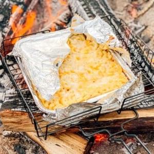 Einfaches Lagerfeuer Mac and Cheese 9 Eine Aluminiumschale mit Lagerfeuer Mac & Cheese zischt auf einem Grill über offenen Flammen, umgeben von Holzscheiten.