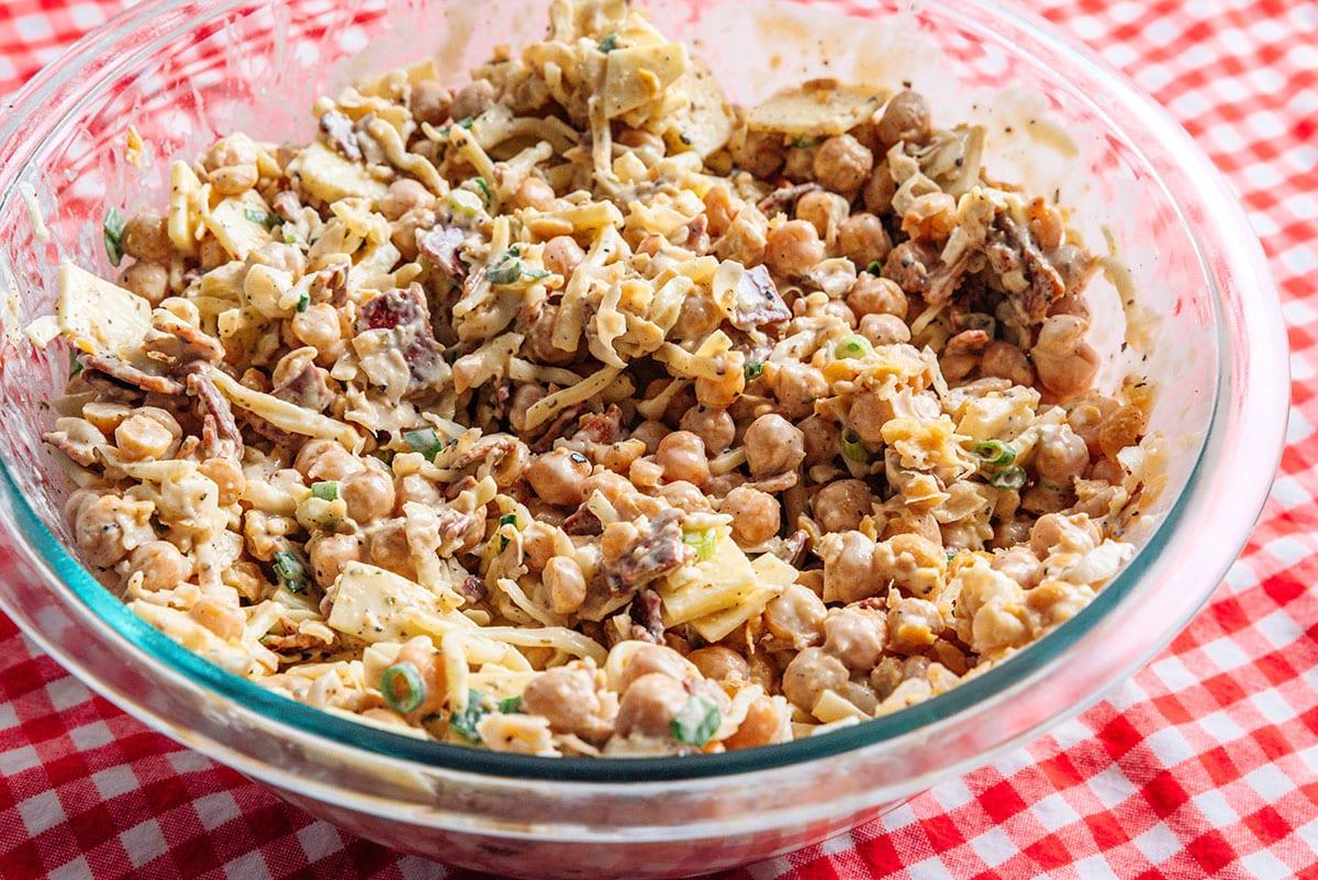 Eine Glasschüssel mit einer Mischung aus Kichererbsen, geriebenem Käse, gewürfeltem Gemüse und einem cremigen Dressing auf einer rot-weiß karierten Tischdecke.