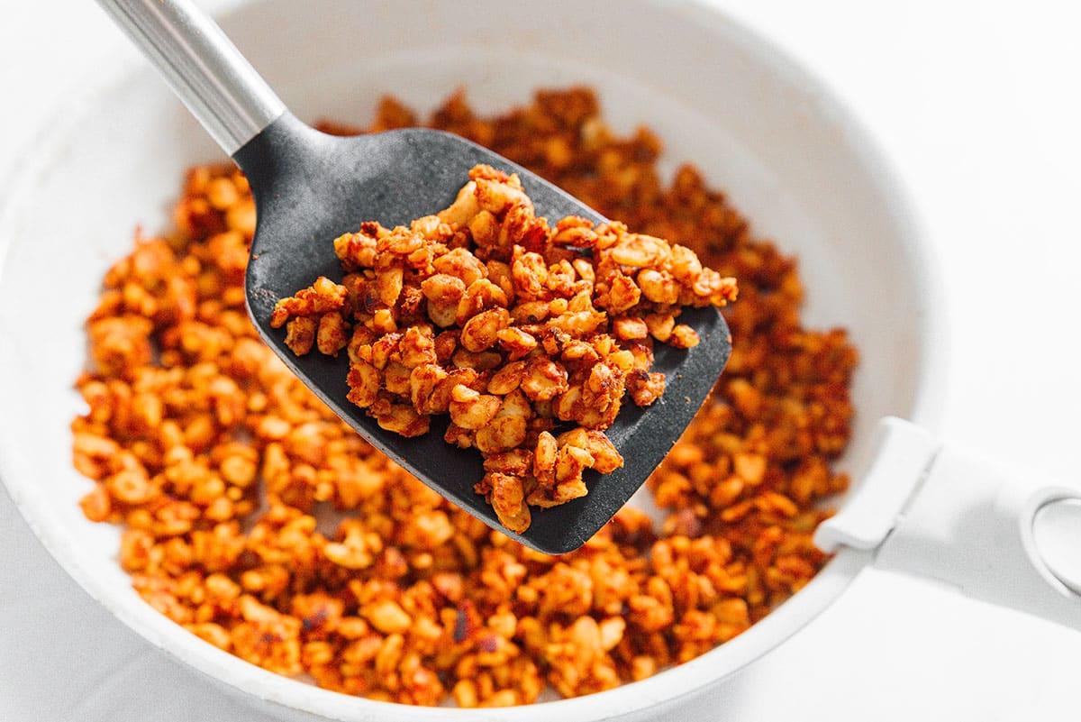 Veganes Tempeh-Taco-Fleisch in einer Pfanne auf weißem Hintergrund, während es mit Gewürzen und Tomatenmark aromatisiert wird.