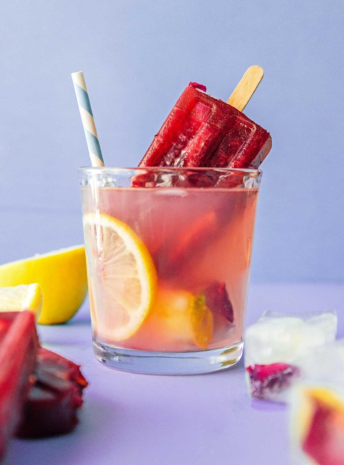 Einfache Lavendel-Zitronenlimonade (Saft oder Slush!) 10 Ein Glas Lavendel-Zitronenlimonade, gefüllt mit essbaren Blüten-Eiswürfeln und Zitronenscheiben, gekrönt mit einem leuchtend roten Hibiskus-Eis am Stiel, auf einem rustikalen Holztisch.