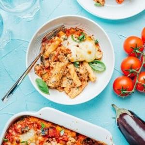 Einfaches vegetarisches Ziti-Auflauf-Rezept (Gemüsereich!) 10 Baked vegetable ziti with cheese and herbs in a white dish, nestled beside plates of the same, fresh tomatoes, eggplant, and empty glasses on a light blue surface.