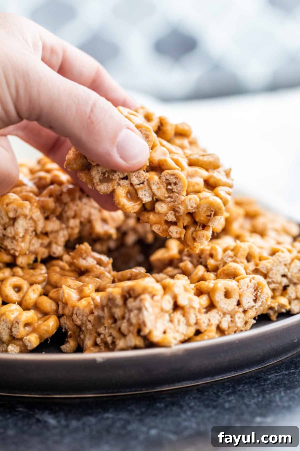 A hand reaching for a peanut butter cheerio bar from a stack on a plate, highlighting the deliciousness and portability.