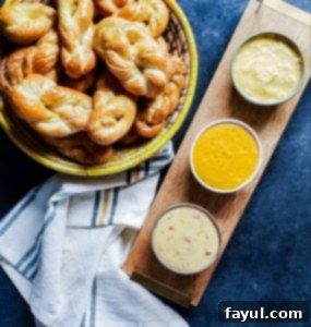 A woven yellow and cream basket with homemade soft pretzel twists. Beside it, a wooden serving tray holds three types of cheese dips on a blue counter.