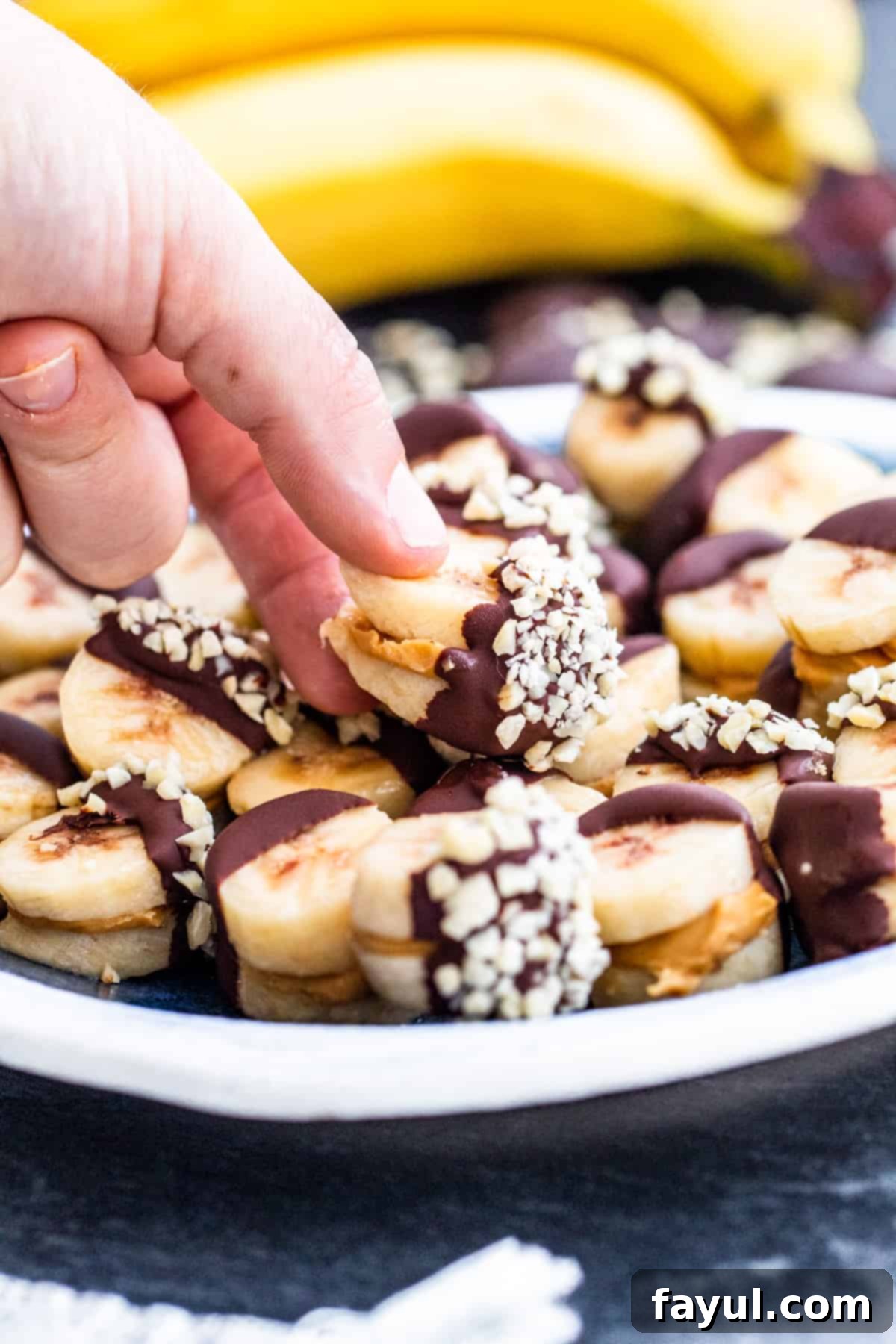 A hand gently picking up one perfect frozen chocolate peanut butter banana bite from a white plate, ready to be enjoyed.