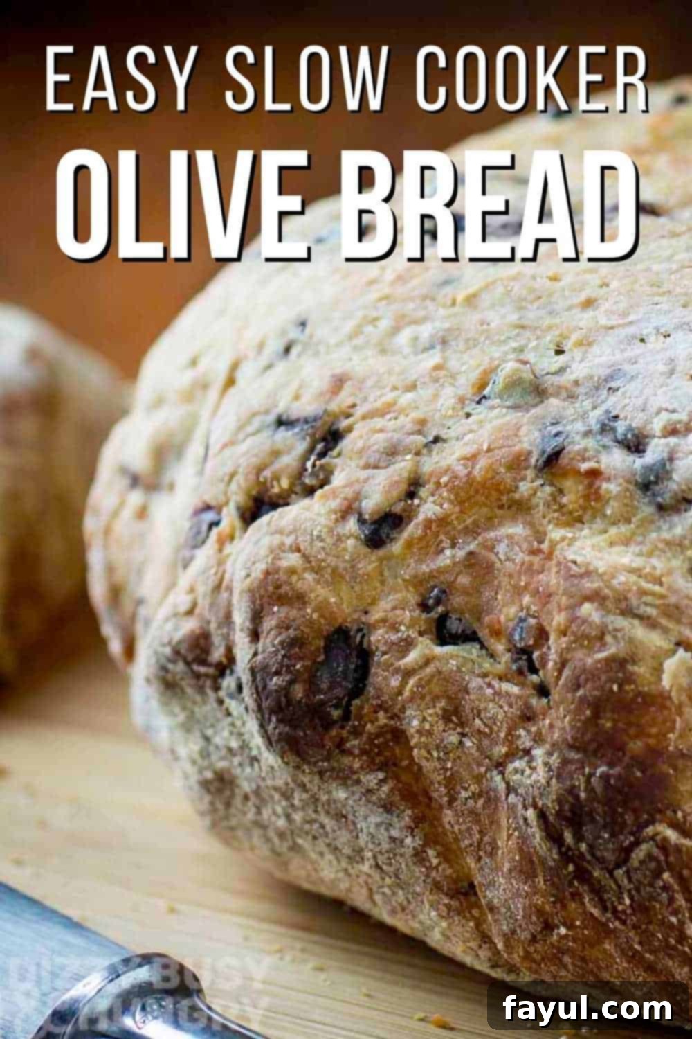 Side view of a whole loaf of golden-brown olive bread on a wooden cutting board, with a butter knife resting beside it. The bread has a beautiful rustic crust and visible olive pieces.