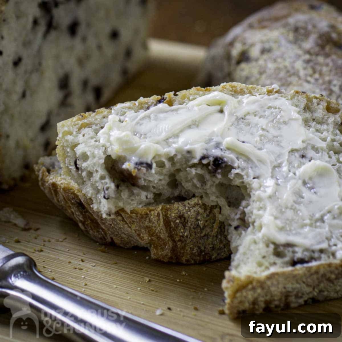 Close-up shot of a thick slice of olive bread, spread with butter, with the blurry whole loaf in the background on a wooden cutting board with a butter knife.