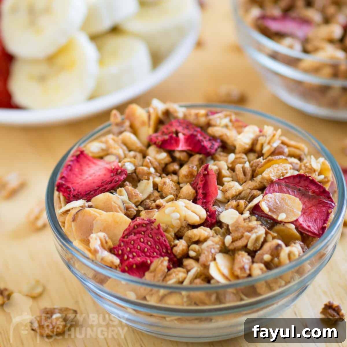 Double Strawberry Granola Delight 6 Side view of a bowl of healthy strawberry granola with dried strawberries, placed on a wooden surface with a backdrop of more granola and freshly sliced fruits.