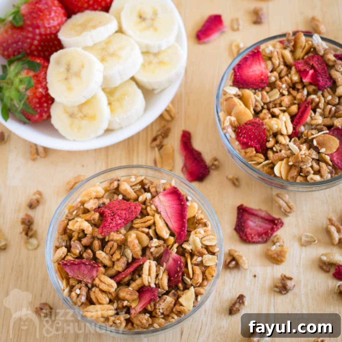 Double Strawberry Granola Delight 7 Overhead shot of two clear bowls filled with homemade granola and dried strawberries, casually sprinkled with more granola on a wooden surface, alongside a bowl of assorted sliced fresh fruit.