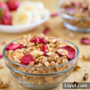 Double Strawberry Granola Delight 8 Side view of healthy homemade strawberry granola in a clear bowl on a wooden surface, with additional granola and sliced fruits in the background.