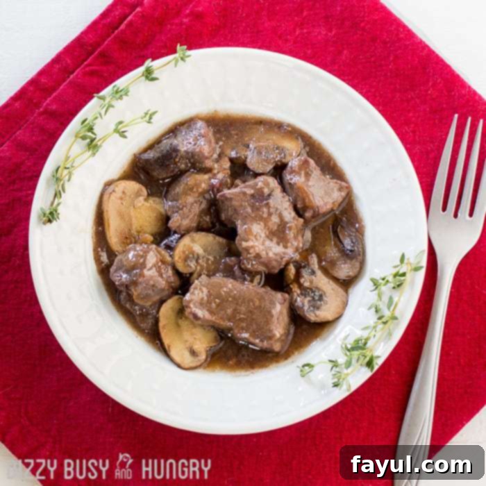 Overhead shot of crock pot beef and mushrooms in a white bowl garnished with herbs on a red cloth with a fork on the side.