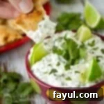 Close up shot of a chip being dipped into cilantro lime dip with sliced limes and cilantro in the background.