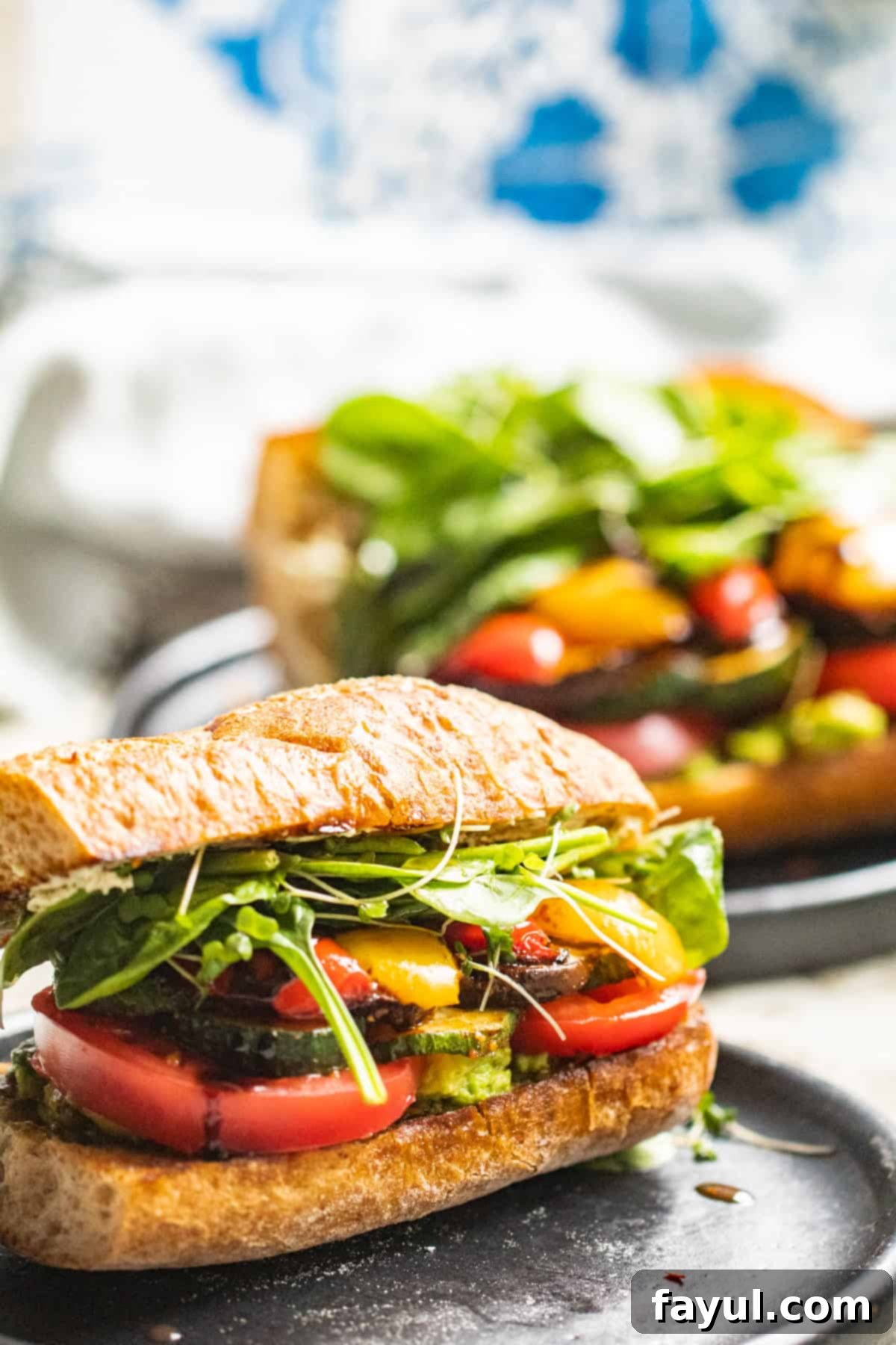 Two roasted vegetable sandwiches on dark gray plates sitting on a white counter.