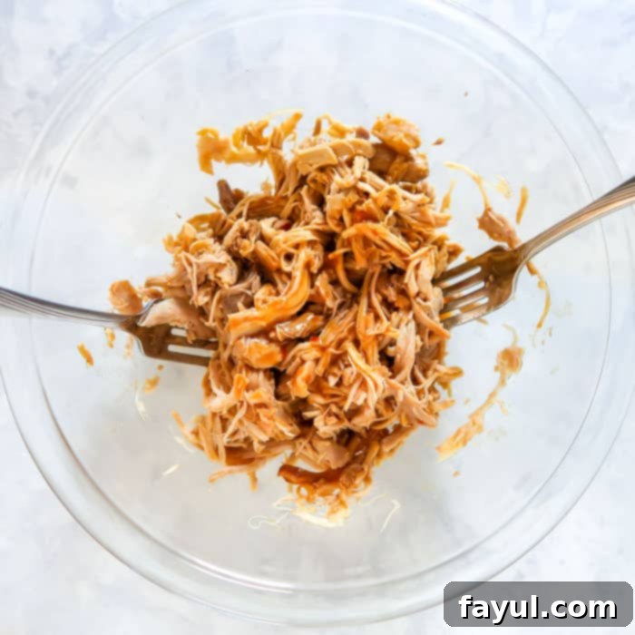 Overhead shot of chicken pulled into stringy bite-sized pieces by two forks in a clear bowl.
