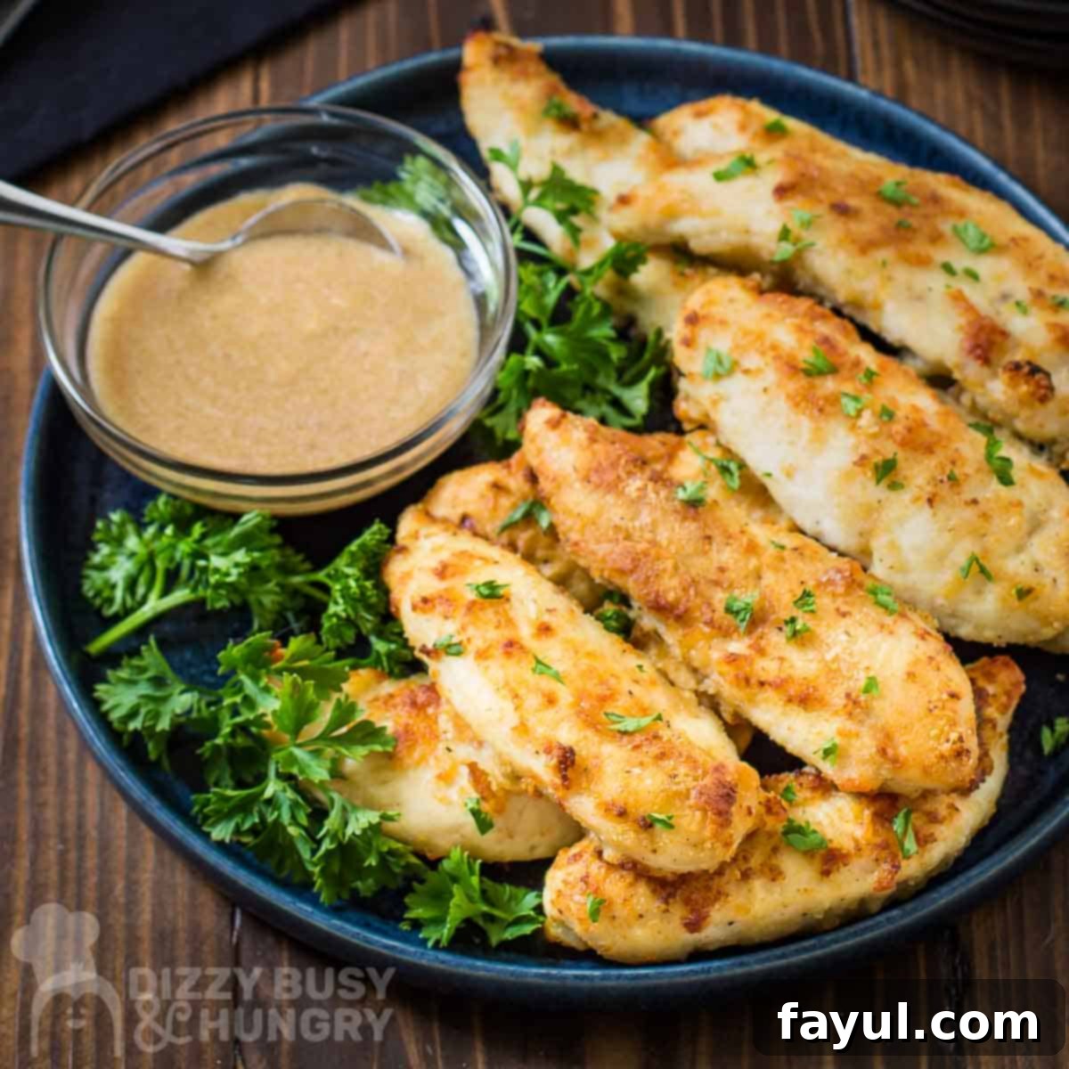 Side shot of perfectly golden pan-fried chicken tenders arranged on a bed of fresh mixed greens on a sleek black plate, with a small clear bowl of savory gravy situated on the side, all set on a warm wooden surface.