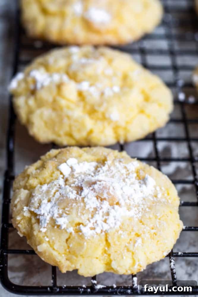 A line of freshly baked Gooey Butter Cake Cookies cooling on a wire rack, showcasing their perfectly cracked tops and delicate powdered sugar coating.