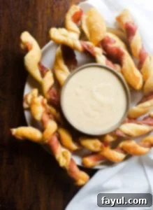 Prosciutto wrapped breadsticks on a white plate with cheese sauce in the middle on a wooden counter.