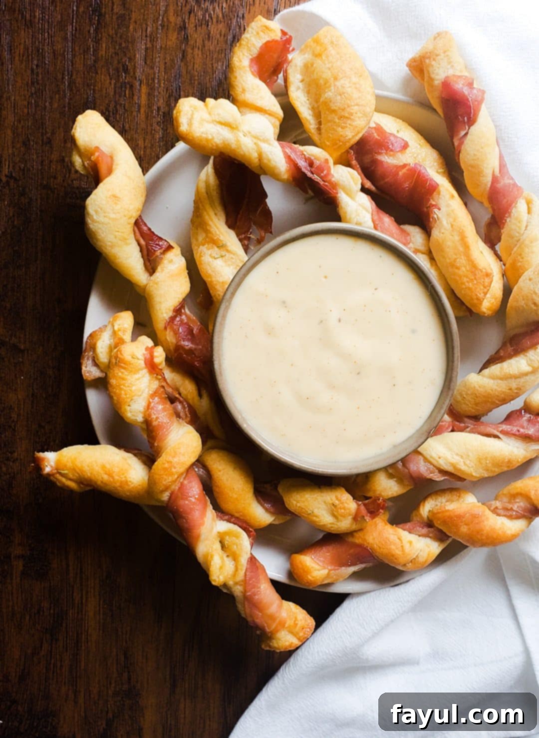 Image for Prosciutto Wrapped Breadsticks with Gouda Cheese Dip recipe. The image is an above shot. It shows a white plate with cheese dip in a gray bowl in the center of the plate. Surrounding the dip on the plate is the twists. The plate is sitting on a wooden table with a white cloth on the right side.