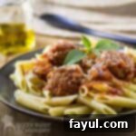 Side shot of delicious meatballs and sauce served over a bed of penne pasta, garnished with fresh basil on a black plate.