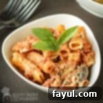 Close-up shot of a white bowl filled with baked ziti, garnished with fresh basil, resting on a rustic brown surface.