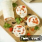 Overhead shot of three perfectly cut pinwheels from a log, arranged on a wooden cutting board, surrounded by sliced olives, fresh herbs, and vibrant red peppers.