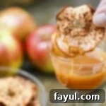 Side close-up shot of a hand dipping a cinnamon apple chip into a glass of caramel sauce, with fresh apples blurred in the background.