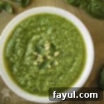 Overhead shot of vibrant broccoli pesto in a white bowl, elegantly garnished with fresh basil and pine nuts, with additional basil sprigs on a wooden cutting board in the background.