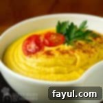 Close-up side shot of creamy hummus, beautifully garnished with halved cherry tomatoes, fresh herbs, and a dusting of paprika, served in a white bowl on a wooden surface.