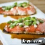 Side shot of two pieces of salmon garnished with green onions, almonds, and sauce on a white square plate.