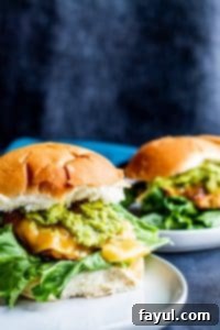 Close-up of a perfectly grilled chicken burger topped with melted Gouda cheese, fresh lettuce, and a generous dollop of homemade guacamole and garlic aioli, served on a toasted bun. A second burger is visible in the background, out of focus.