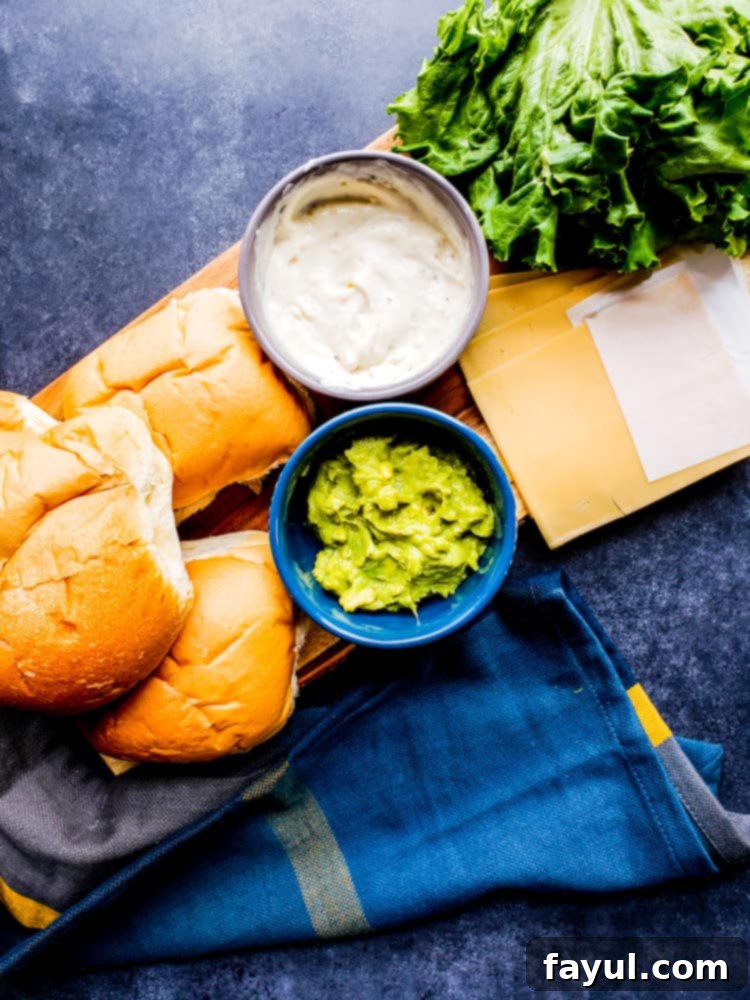 Overhead shot of all fresh ingredients for grilled chicken burgers and garlic aioli laid out on a wooden cutting board, including ground chicken, spices, egg, milk, breadcrumbs, mayo, garlic, lemons, and buns.