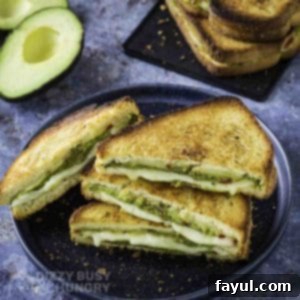 Side shot of multiple triangle halves of avocado grilled cheese on a black plate with more on a black plate and an avocado in the background.