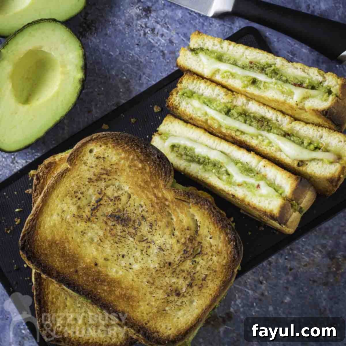 Overhead shot of full and halved avocado grilled cheese on a black rectangle plate on a blue marbled surface with a knife and halved avocados on the side.