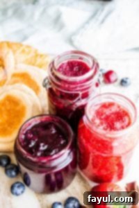 Strawberry, blueberry, and cherry jam in glass containers with fruit around them.