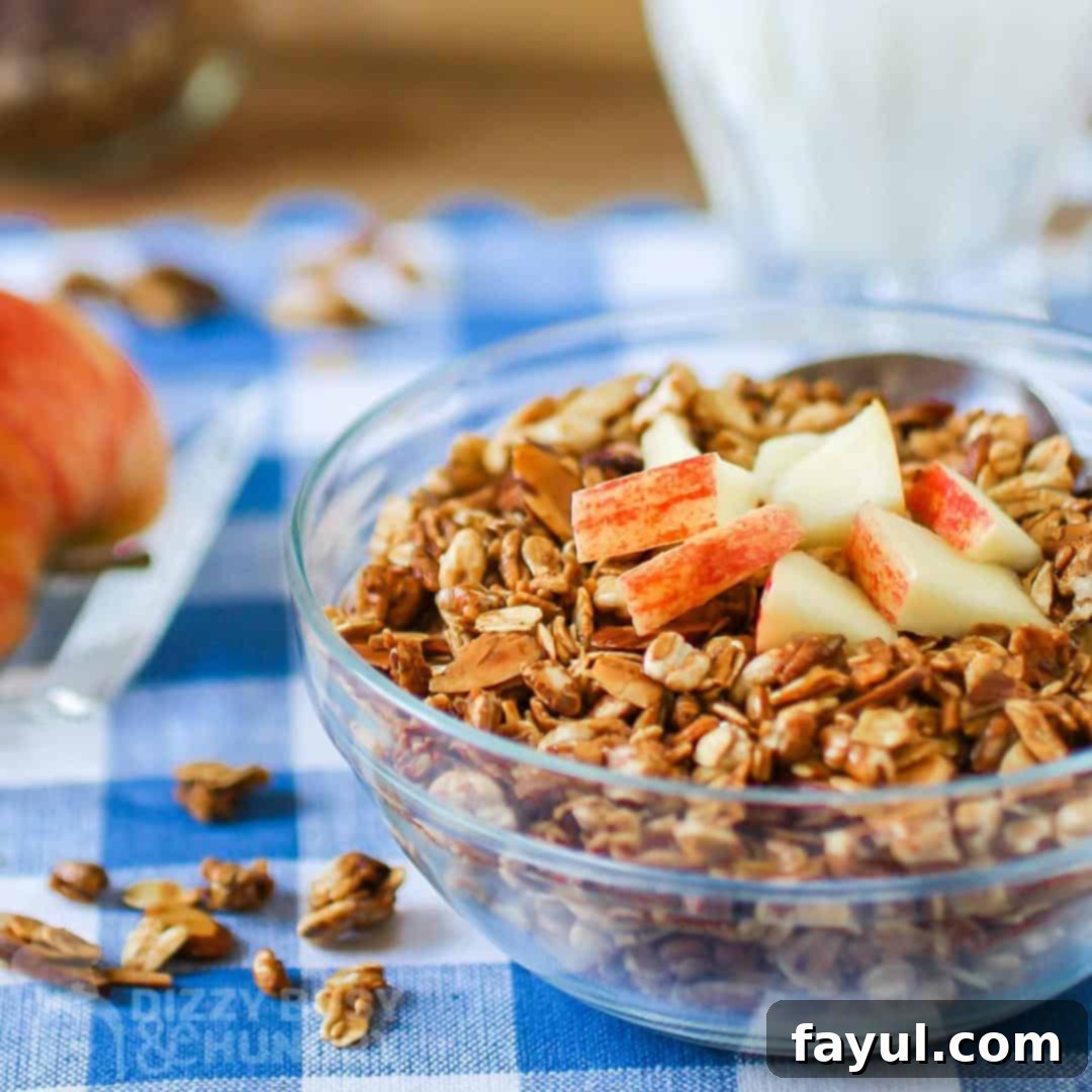 Bowl of granola topped with chopped apples, on a blue checked napkin with a pitcher of milk in the background.