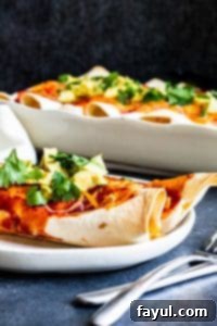 Shot of two veggie enchiladas on a white plate with silver fork and knife on the side. A white baking dish with more in the background.