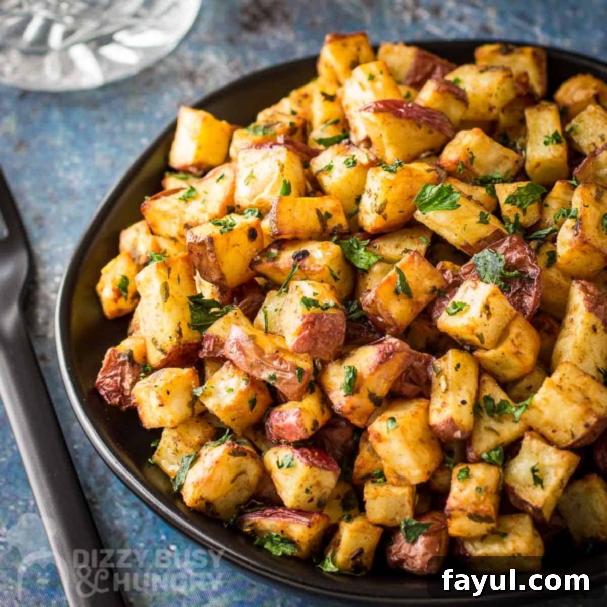 Side close up shot of perfectly golden and crispy air fryer breakfast potatoes served on a black plate, resting on a blue speckled surface with black utensils on the side, highlighting their delicious texture.