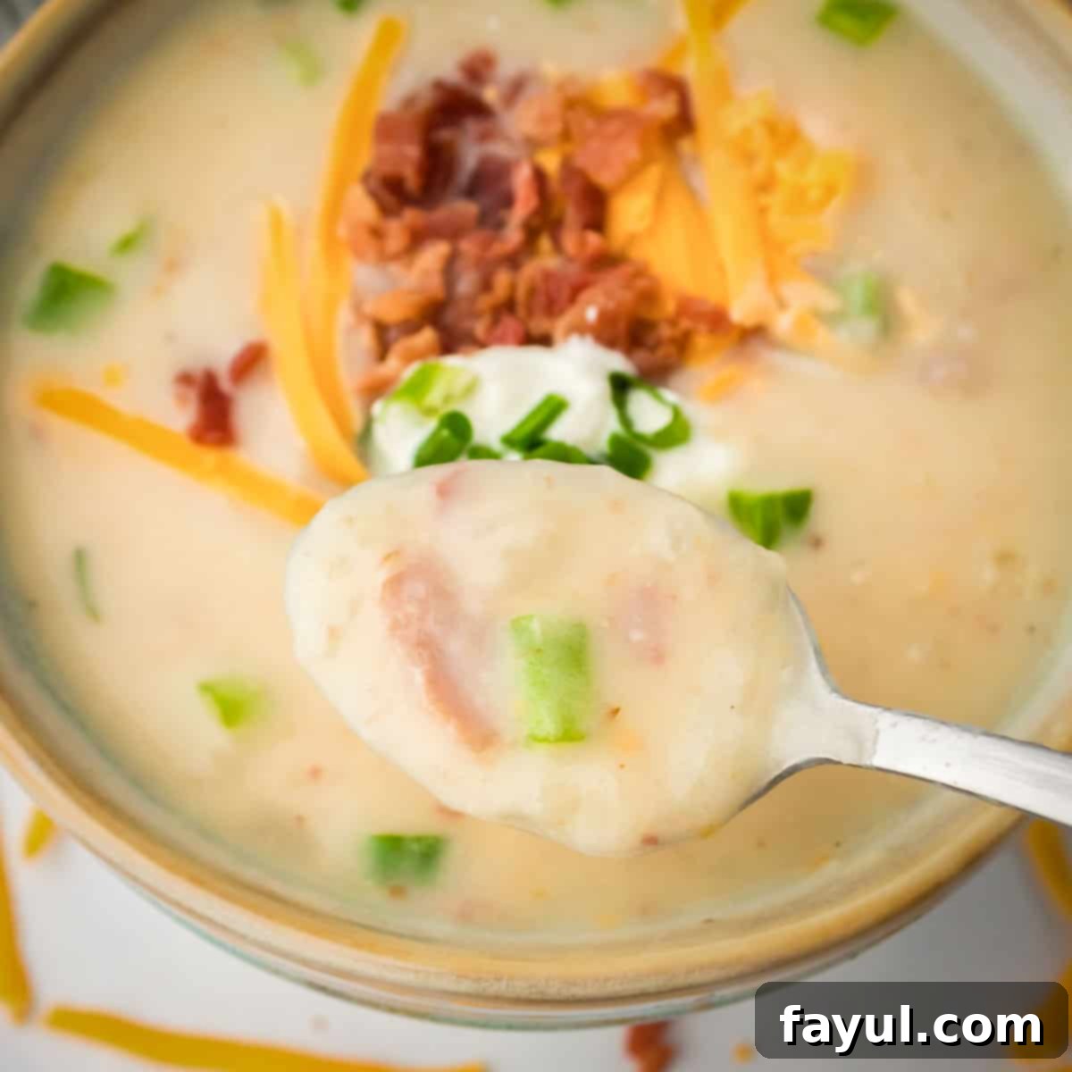 Overhead shot of a spoon holding some potato soup garnished with bacon, cheese, and chives in a green bowl.