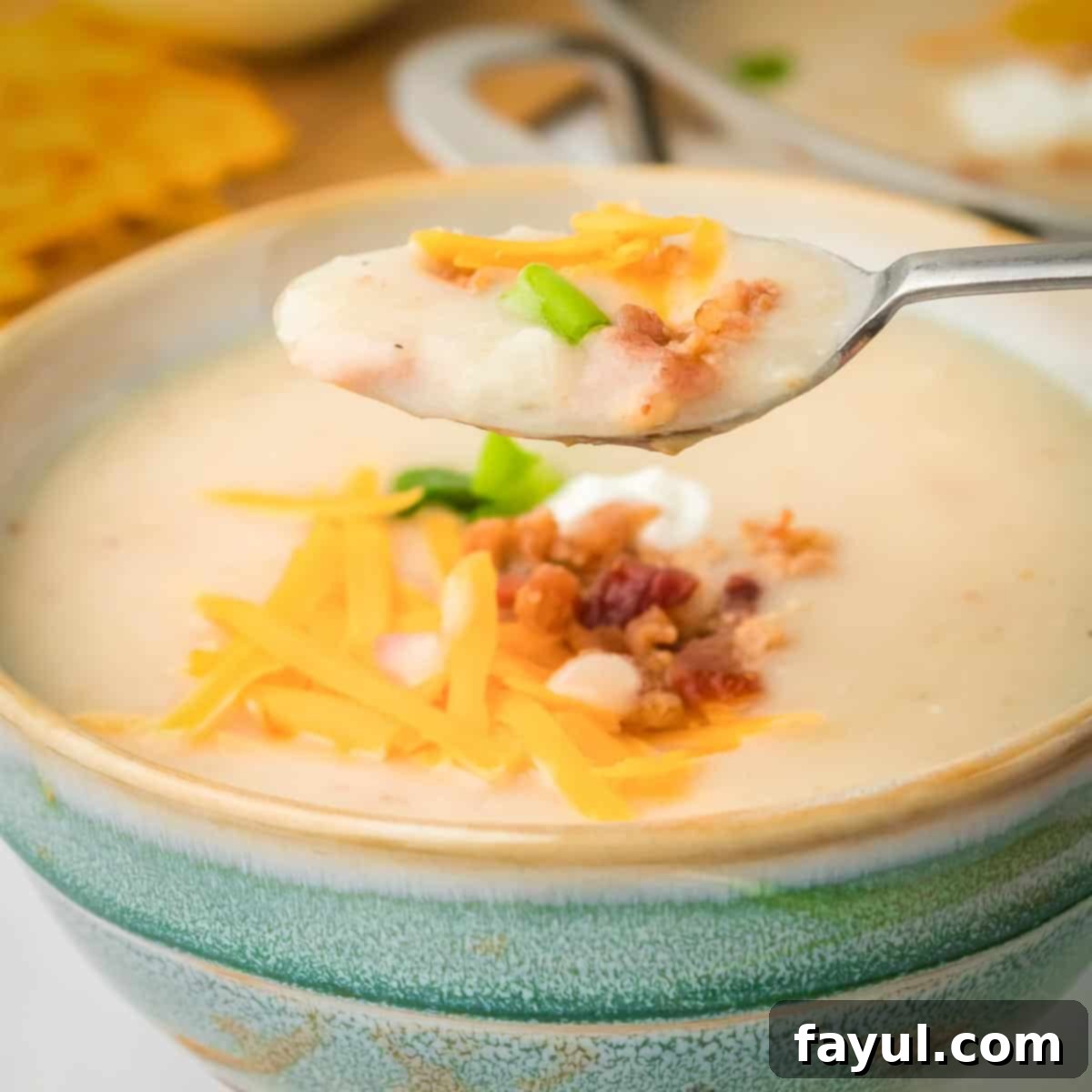 Side close up shot of a spoon holding some potato soup garnished with bacon, cheese, and chives in a green bowl.