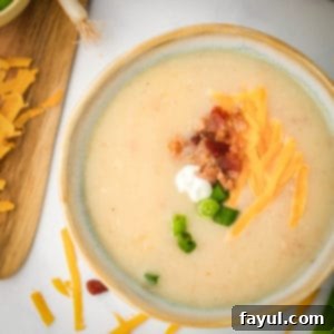 Overhead shot of potato soup garnished with shredded cheese, bacon, chives, and sour cream on a white surface.
