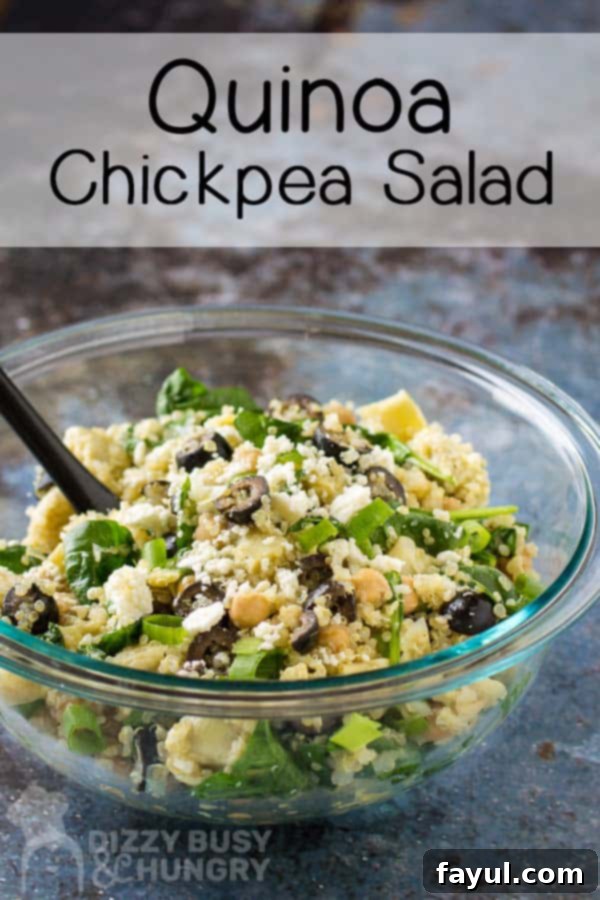 Side close-up view of a freshly made Quinoa Chickpea Salad in a clear bowl with a black spoon, set against a subtle grey background. The salad looks vibrant and inviting with clearly visible artichokes, chickpeas, and feta.