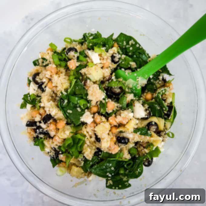 The previous quinoa mixture, now vibrant with the addition of crumbled feta cheese, freshly chopped scallions, and tender baby spinach, all being gently folded together in a clear glass bowl with a green spatula.