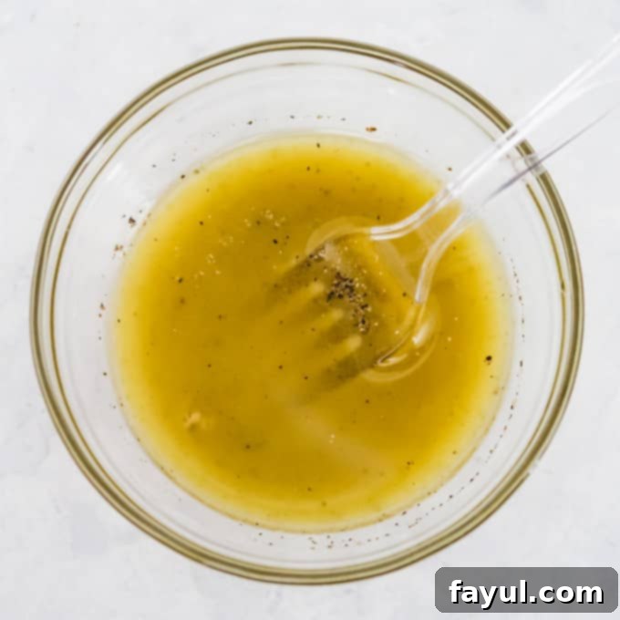 Overhead shot of dressing ingredients – including golden olive oil, vibrant lime juice, aromatic garlic paste, and a sprinkle of salt and pepper – perfectly mixed together in a clear glass bowl with a clear fork, set against a clean white background.