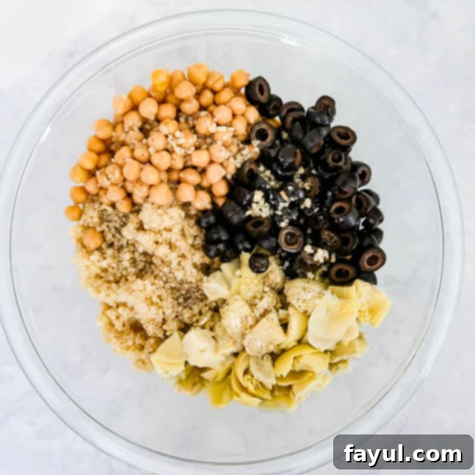 Close-up shot of plump chickpeas, glossy sliced black olives, fluffy cooked quinoa, and tender chopped artichoke hearts, along with the vibrant dressing, all being mixed together in a clear glass bowl.