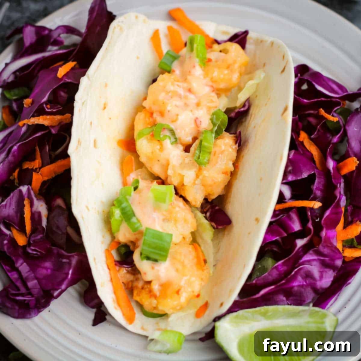 Overhead close up view of a shrimp taco on a bed of purple cabbage and shredded carrots with a lime wedge on the side on a round white plate.