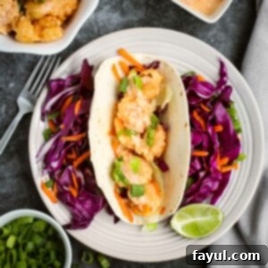 Overhead shot of one shrimp taco with shredded carrots and purple cabbage surrounding it and a lime wedge on the side of the white plate. On the black surface surrounding, there is more shrimp, sauce, and chopped green onions in white bowls as well as a fork and grey cloth.
