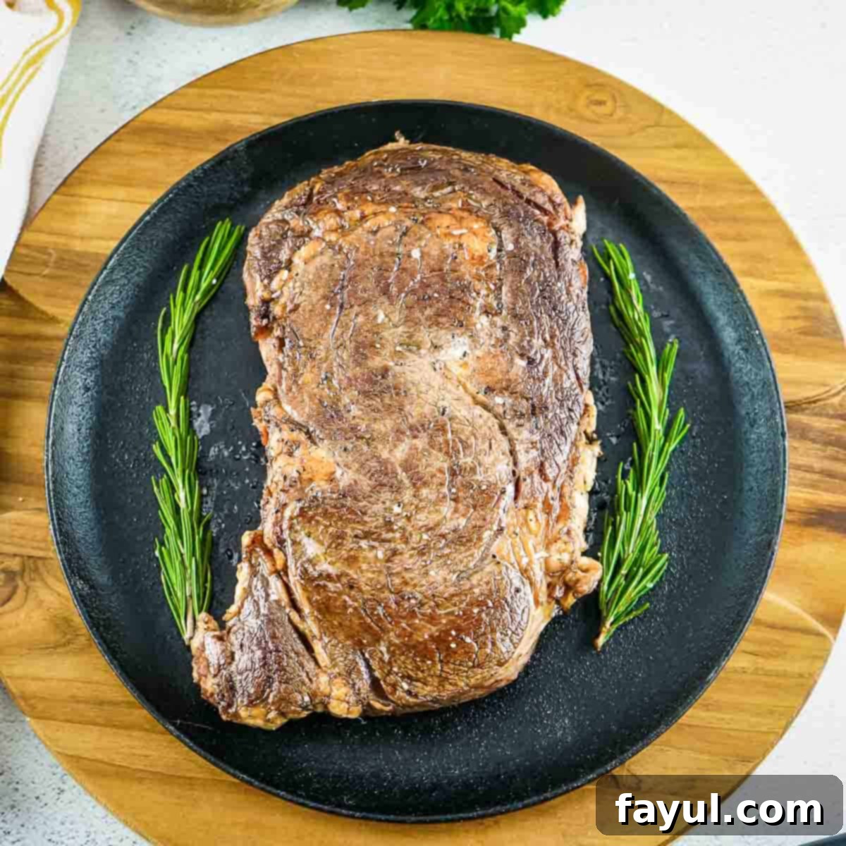 Overhead shot of whole ribeye steak with garnishes of rosemary on the side all on a black plate on a round wooden surface.